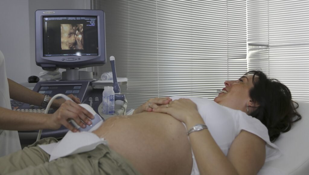 A side view of a smiling, pregnant woman lying on an examination bed, receiving a diagnostic ultrasound. A medical professional, whose hands are visible in the foreground, applies the transducer to the woman's abdomen with gel. On the monitor to the left, a 3D image of the fetus is clearly displayed. The background features gray horizontal window blinds.
